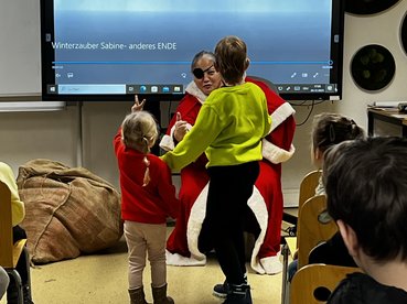 Eine als Weihnachtsfrau verkleidete Mitarbeiterin sitzt auf einem Sessel. Vor ihr stehen 2 Kinder.