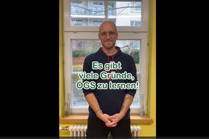 Ein Kursteilnehmer steht mit verschränkten Händen vor einem Fenster. Vor ihm ist ein grün-weißter Text: "Es gibt viele Gründe, ÖGS zu lernen"
