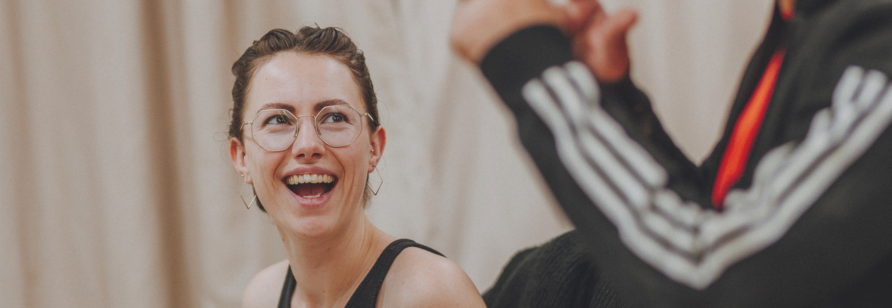 Junge Frau mit Brille lacht jemanden an, der nur halb im Bild zu sehen ist.