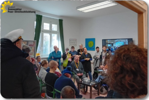 Zu sehen sind Mitarbeitende und Besuchende von der Agentur Sonnenklar. Sie sitzen mit aufmerksamer Mimik rund um einen kleinen Tisch.