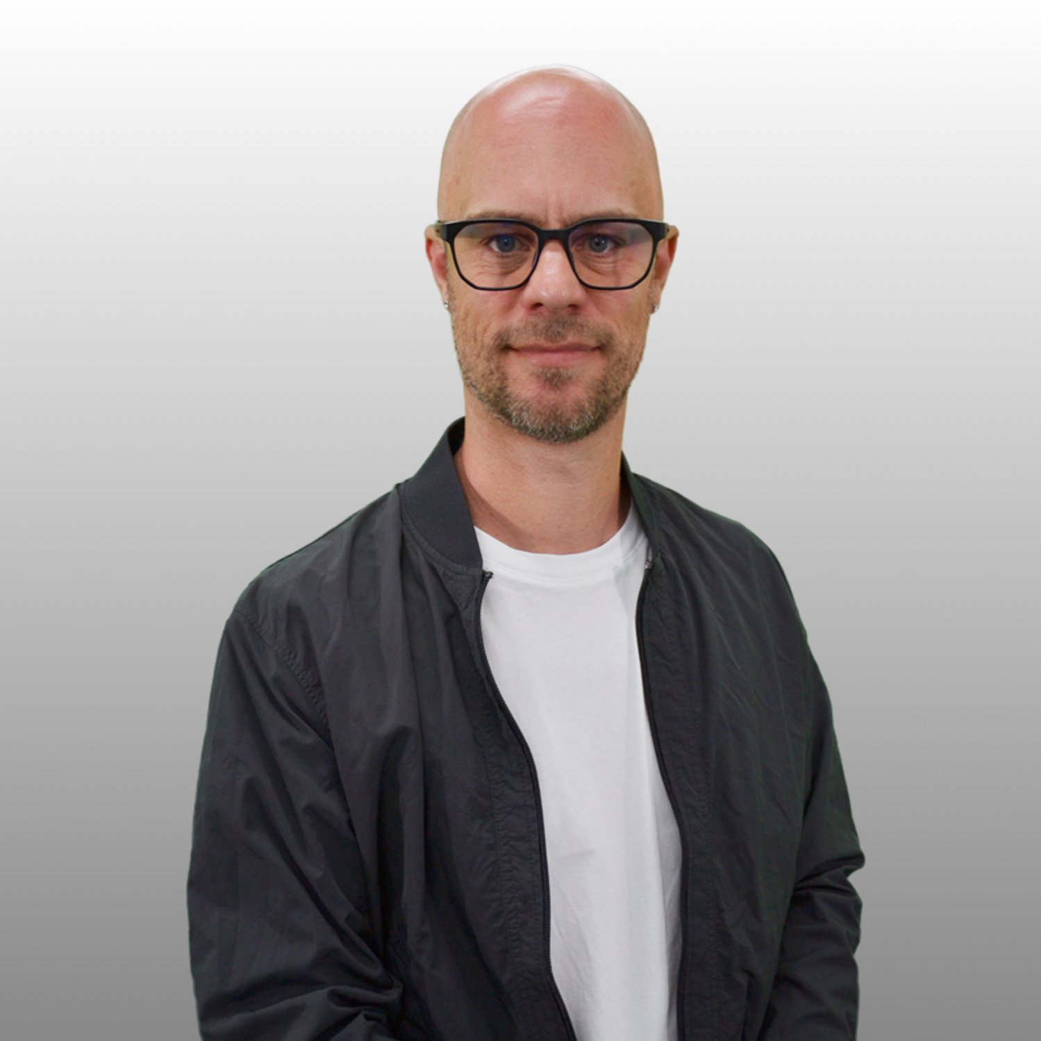 Foto von Thorben Pachler vor grauem Hintergrund: Mann, Mitte dreißig, mit Brille, Glatze und blondem kurzem Vollbart trägt ein weißes Shirt mit blauem, offenen Hemd darüber.