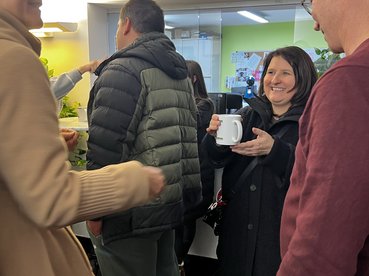 Elicabeth Flack vom Fördergeber Sozialministeriumservice steht in einer Runde mit weiteren Persone. Sie befindet sich in der Cafeteria des equalizent und hält eine Tasse mit Punsch in der Hand.n