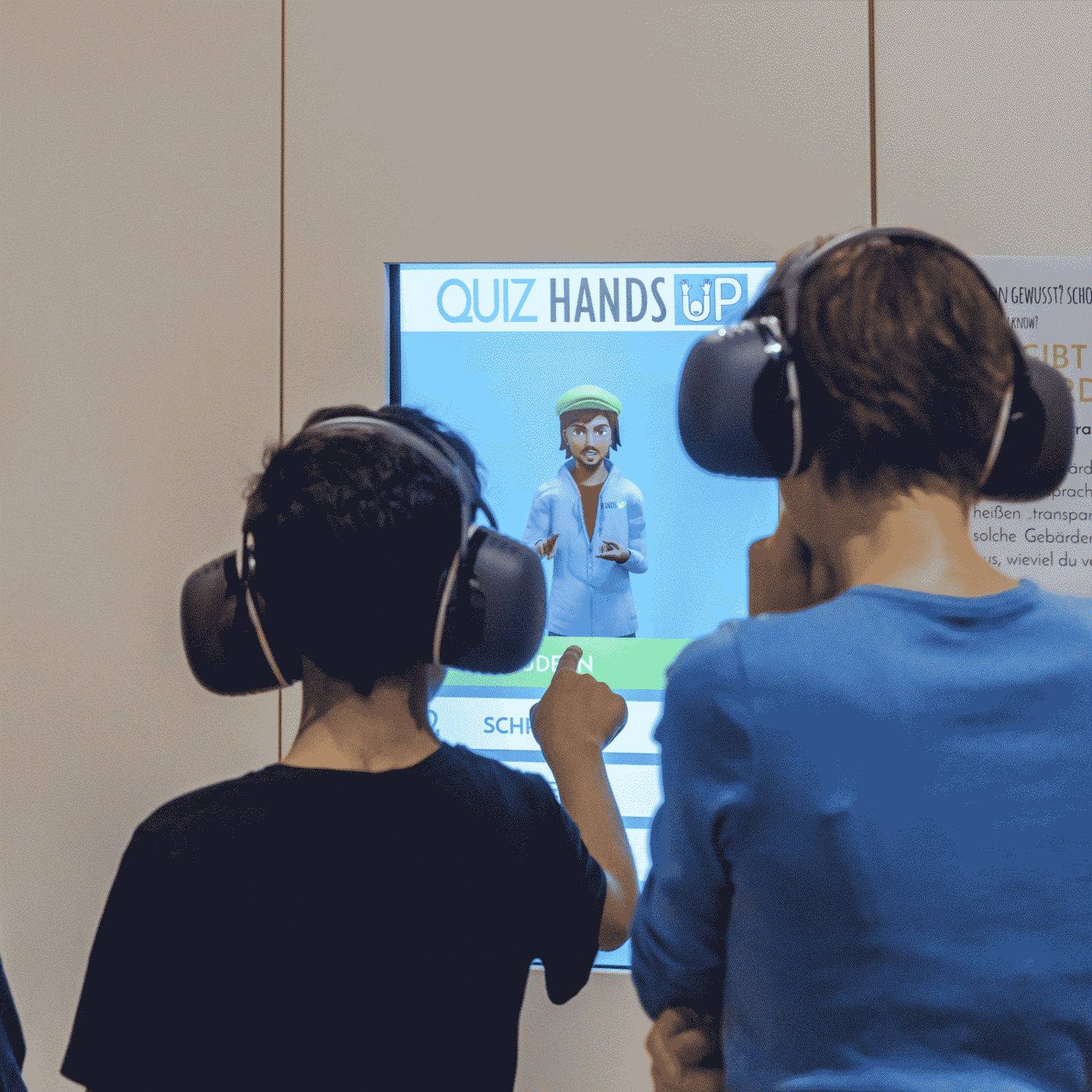 Zwei Kinder stehen mit dem Rücken zur Kamera. Beide tragen Kopfhörer. Vor ihnen ist ein Touchscreen mit einem Avatar. Ein Kind tippt mit dem Finger auf den Bildschirm.