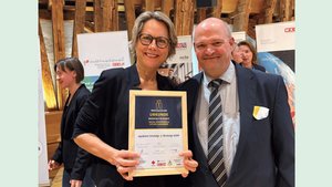 Zu sehen sind eine Frau mit Brille und blöden, kürzeren Haaren, sowie ein Mann mit Halbglatze. Beide lächeln und tragen elegante Kleidung. Sie hält eine Urkunde des Trigos-Awards in der Hand. Im Hintergrund sieht man verschiedene Rollups und ein paar Menschen.