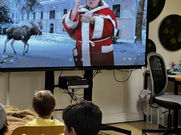 zu sehen ist ein Bildschirm mit einem Standbild des Weihnachtsvideos. Das Bild zeigt das equalizent-Haus im Schnee mit einem Elch und einer als Weihnachtsfrau verkleideten Mitarbeiterin. Das Bild ist KI-generiert. Im Vordergrund sind Kinder von hinten zu sehen, die sich den film anschauen.