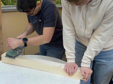 2 junge Männer schleifen mit einer Schleifmaschine ein Stück Holz