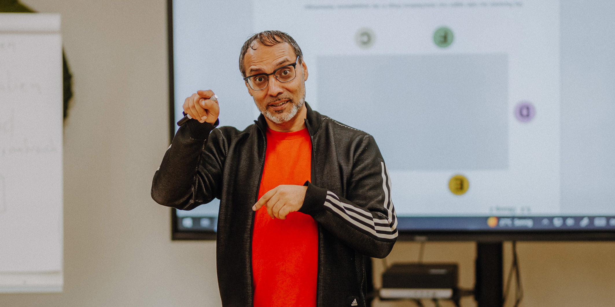 Man mit rotem Shirt und schwarzer Trainingsjacke steht vor einem Smartboard