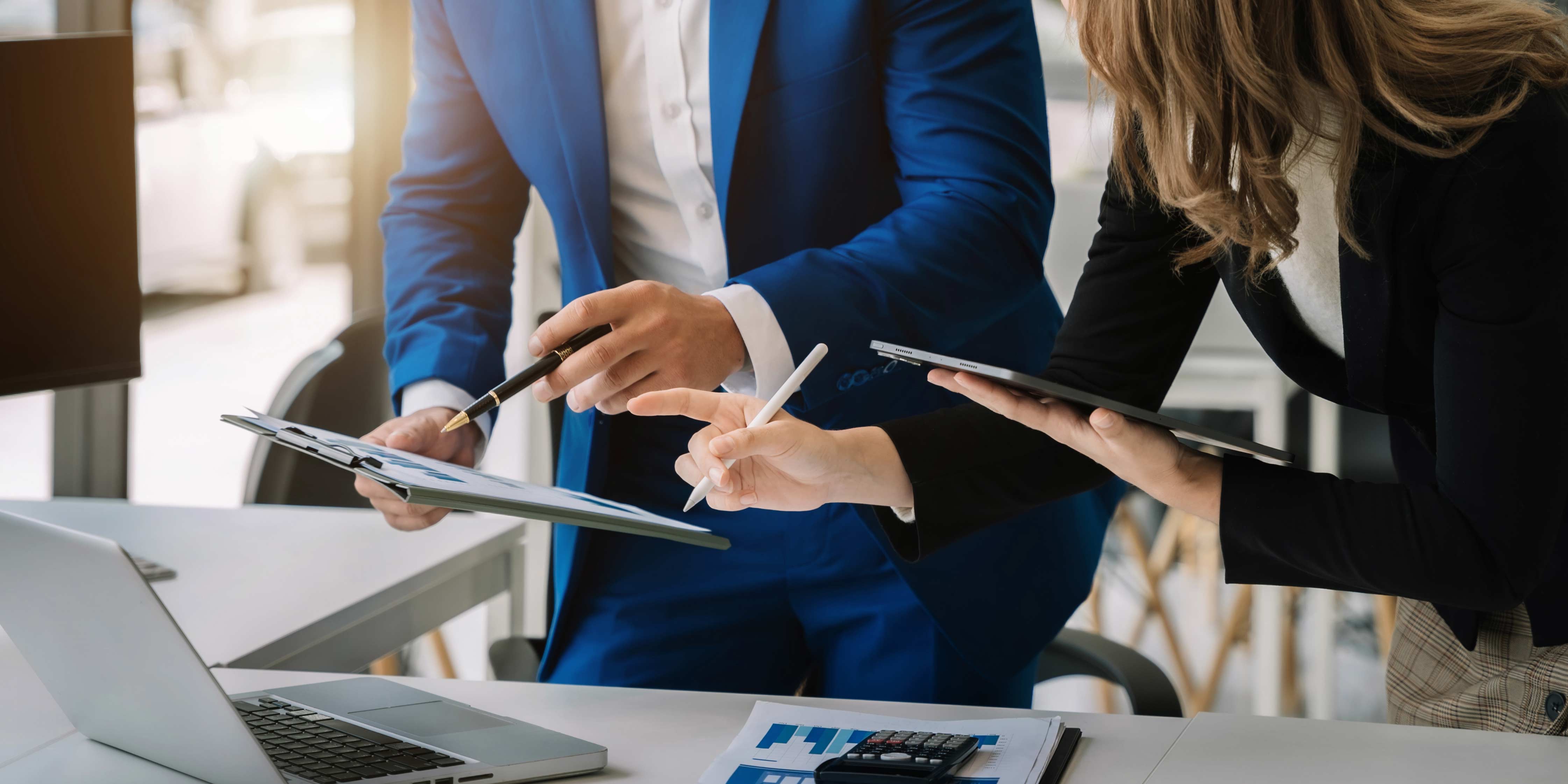 Zwei GEschäftsleute mit Tablet in der Hand und vor einem Laptop stehend diskutieren Businessangelegenheiten