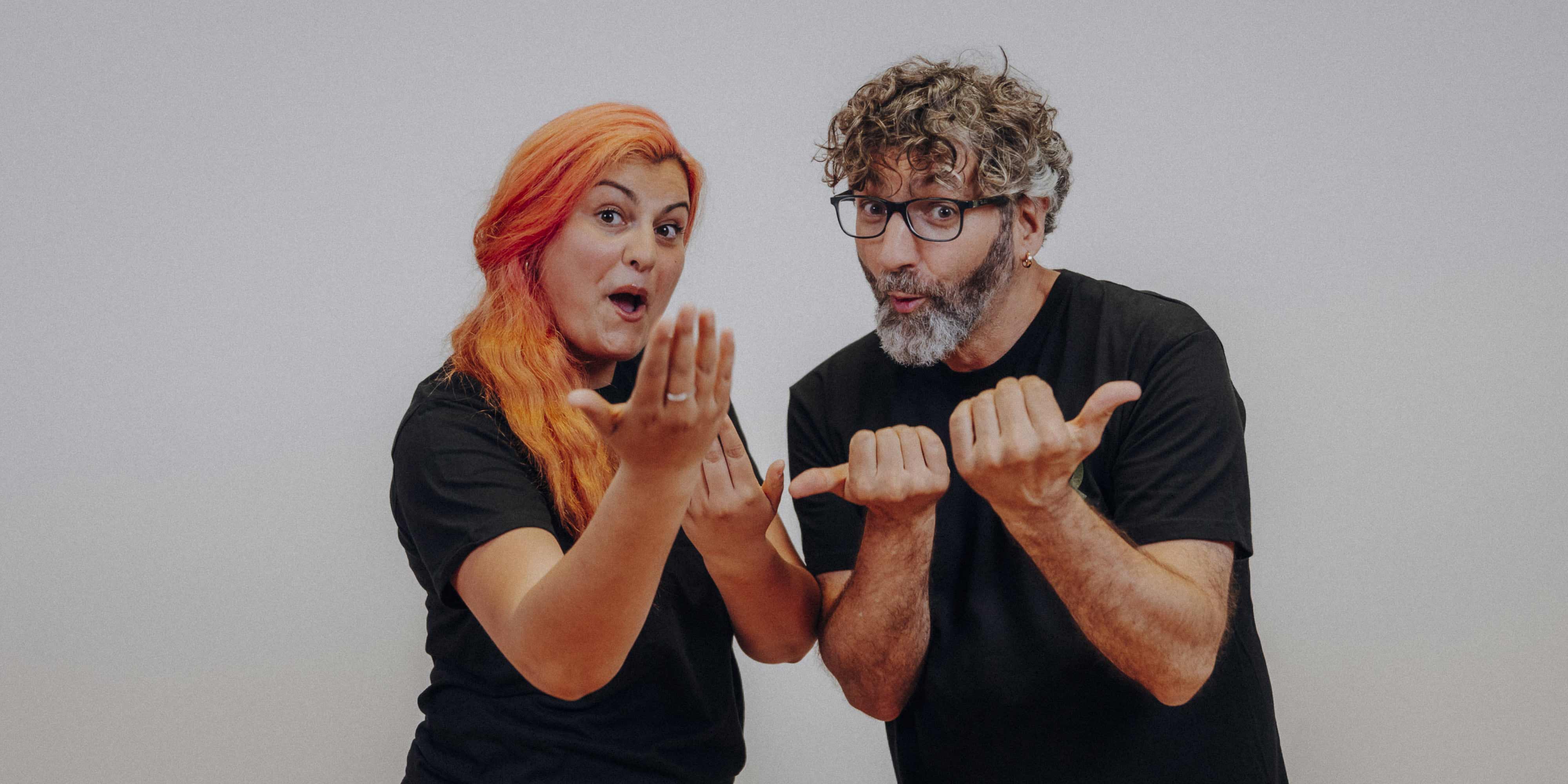 Eine Frau mit orangen Haaren und ein Mann mit grauen Locken, beide in schwarzem T-Shirt gebärden mit beiden Händen "komm!"